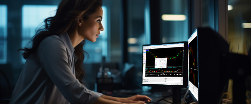 A woman working at a desk with dual monitors, each showing different stock market trend graphs.