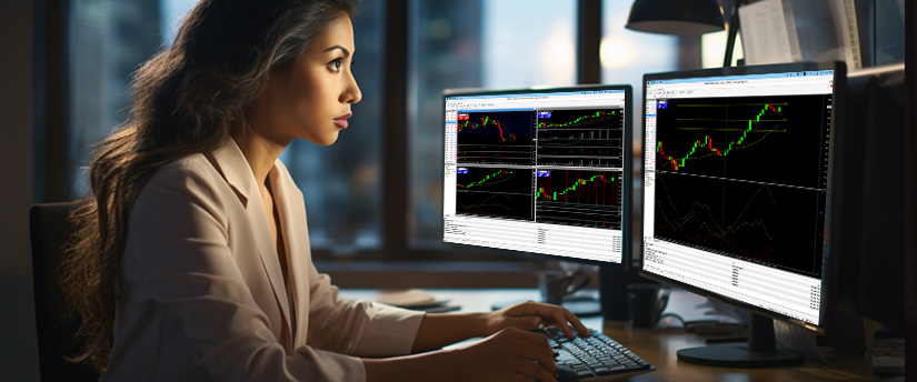 A woman at a desk with two monitors displaying various stock market graphs, focused on forex trading.
